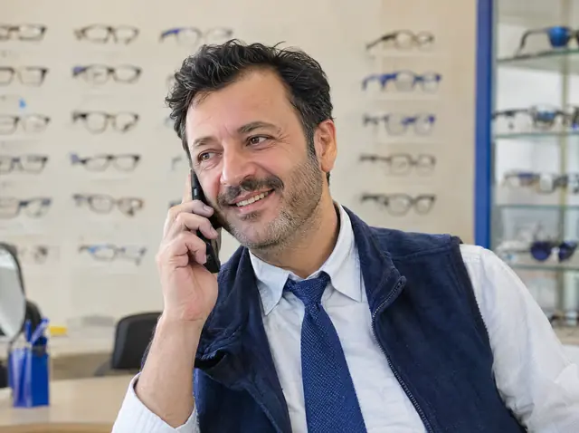 Gözlük raflarının önünde telefonda müşterisine cevap veren, gülümseyen bir optisyen.